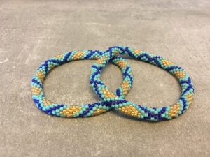 Roll-on armbånd som passer til næsten alle kvinder. Kvindegrupper i Nepal, Kathmandu Valley, udstyres med glasperler og bomuldsgarn, så de i deres fritid kan hækler disse smukke perlearmbånd. Den ekstra indtægt går bl.a. til at sende kvindernes børn i skole. Hvert armbånd er et unikt resultat af kvindernes håndarbejde og varierer i farvekombinationer og mønstre.